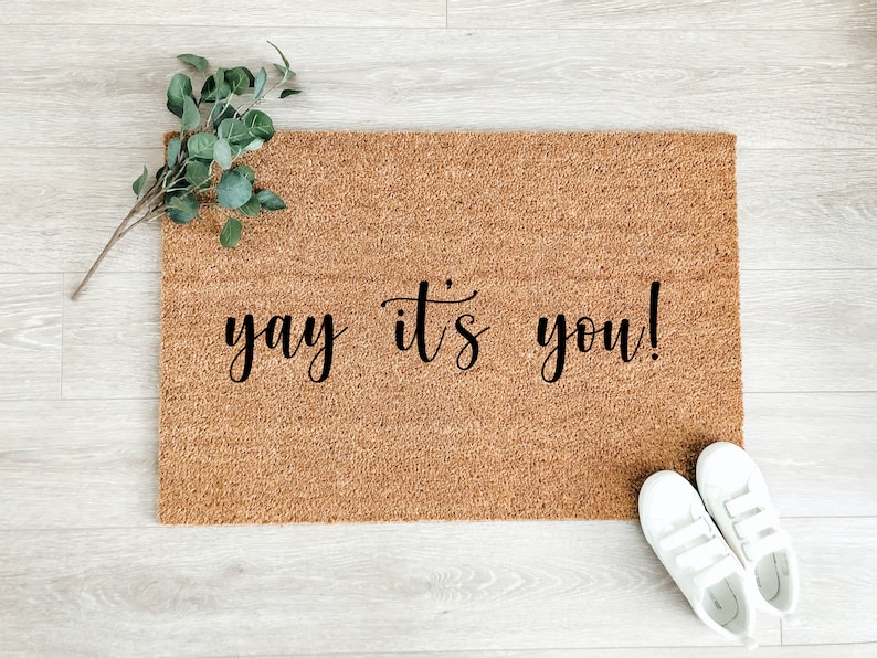 May include: A rectangular coir doormat with the phrase "yay it's you!" in black script. A sprig of greenery and a pair of white sneakers are placed near the mat. The mat is a natural brown color.