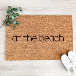 May include: A brown coir doormat with the text "at the beach" in black lettering. Two white canvas shoes are on the floor next to the doormat.