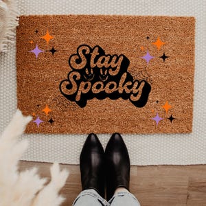 May include: A brown coir doormat with the text "Stay Spooky" in black with a spiderweb design and orange and purple stars.