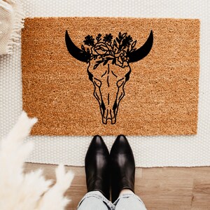May include: A brown coir doormat with a black silhouette of a cow skull with floral accents.