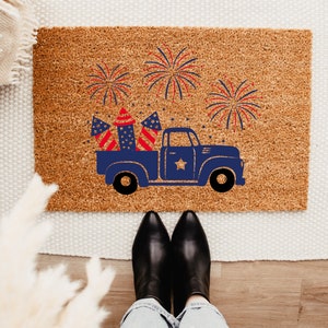 May include: A brown doormat with a blue pickup truck carrying fireworks and American flags. The truck is on a brown doormat with a white background.