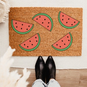 May include: A brown doormat with five watermelon slices printed on it. The watermelon slices are red with black seeds and green outlines. The doormat is on a white rug.