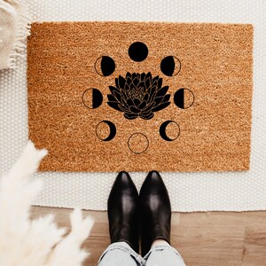 May include: A rectangular doormat made of natural coir fiber. The mat features a black lotus flower design surrounded by phases of the moon. The mat is placed on a white rug and wooden floor.