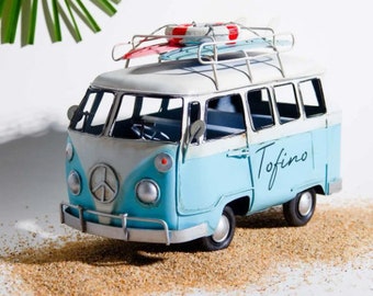 Może przedstawiać: Niebiesko-biały, vintage'owy van z zabawki z deską surfingową i kołem ratunkowym na dachu. Van ma napis "Tofino" na boku.