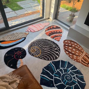 May include: A collection of seven tufted rugs shaped like seashells in various colours and patterns, including black, white, orange, blue, and brown. Each rug features distinct spiral or elongated shell designs with textured surfaces.