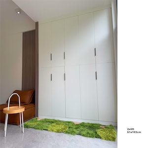 Könnte beinhalten: Ein großer, cremefarbener Kleiderschrank mit schwarzen Griffen. Ein grüner, strukturierter Teppich, 61x183cm, liegt auf dem Boden. Ein kleiner, runder Holztisch mit weißem Rahmen und ein braunes Ledersofa befinden sich links.