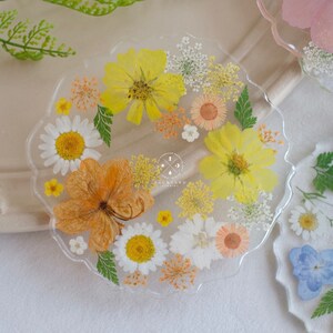 May include: A clear resin coaster with a scalloped edge features pressed flowers in shades of yellow, orange, and white. The coaster has the text "BACKYARD FLOWERS" in the center.