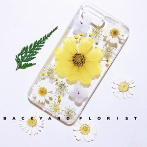 Peut inclure: Une coque de t&eacute;l&eacute;phone transparente avec des fleurs press&eacute;es. La coque pr&eacute;sente une grande fleur jaune entour&eacute;e de fleurs blanches et violettes. Le texte "BACKYARD FLORIST" est imprim&eacute; sur le fond blanc.