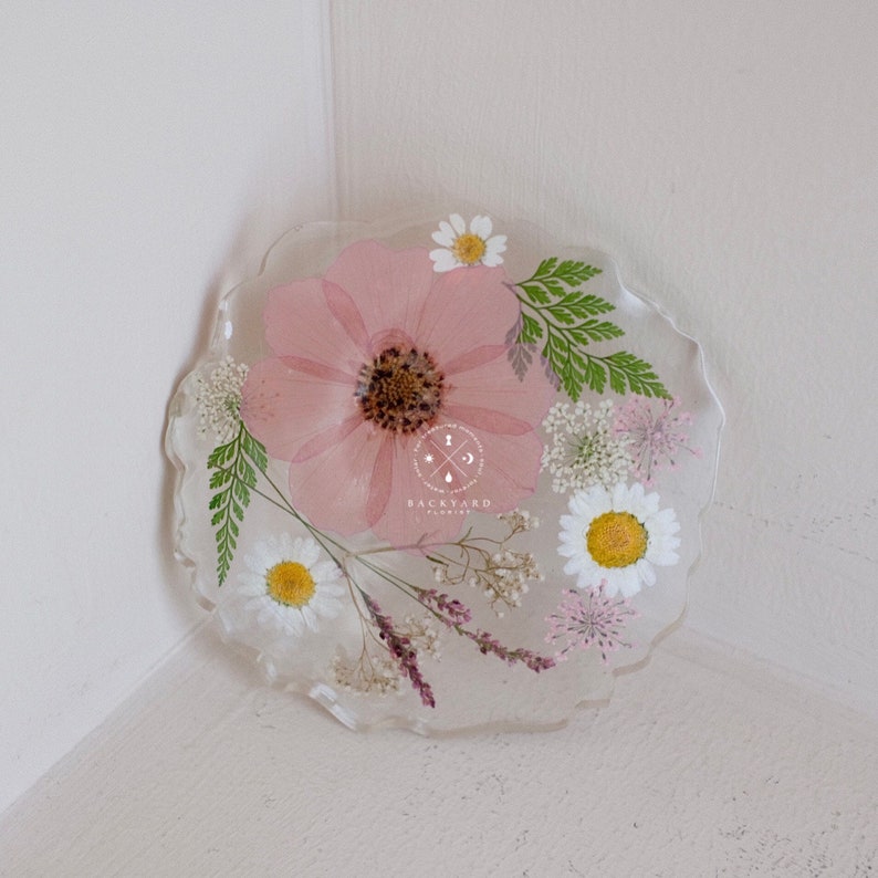May include: A clear resin coaster with pressed pink and white flowers. The coaster features a fern, small white flowers, and a larger pink flower with a yellow center. The text "BACKYARD FLORISTRY" is visible in the center of the coaster.