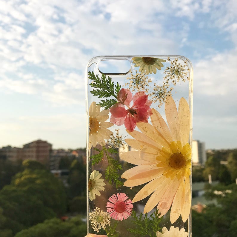 May include: A clear phone case with pressed flowers. The flowers are pink, white, and yellow. The case has a clear background, allowing the flowers to be seen clearly.
