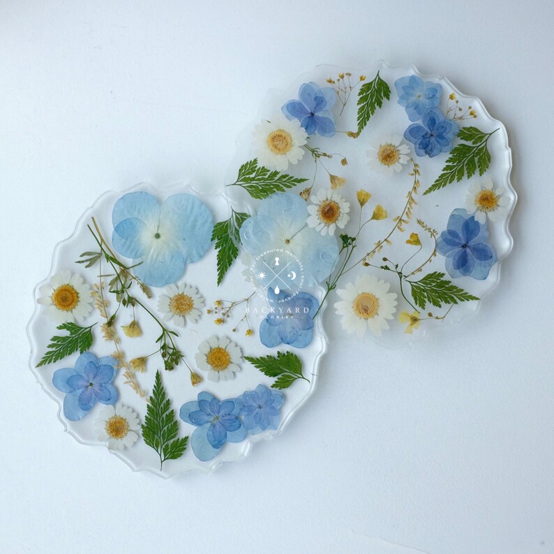 May include: Two round, clear resin coasters with pressed blue and white flowers. The coasters have a scalloped edge and are perfect for adding a touch of nature to your home decor.