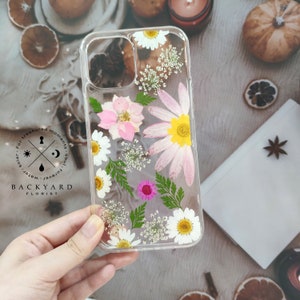 May include: A clear phone case featuring pressed flowers and greenery. The case has a black logo that reads "BACKYARD FLORIST" with the text "For treasured moments. Solar - Water - Forever Soul."