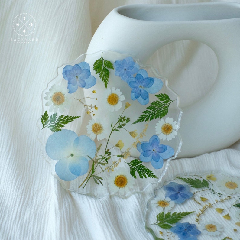 May include: Two round, clear resin coasters with a scalloped edge. The coasters feature pressed flowers and leaves in shades of blue and white. The coasters are on a white fabric surface.