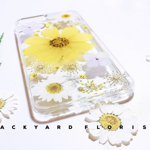 Peut inclure: Une coque de t&eacute;l&eacute;phone transparente avec des fleurs press&eacute;es. La coque pr&eacute;sente une grande fleur jaune avec des p&eacute;tales blancs et des fleurs blanches plus petites. La coque est &eacute;tiquet&eacute;e "BACKYARD FLORIST".