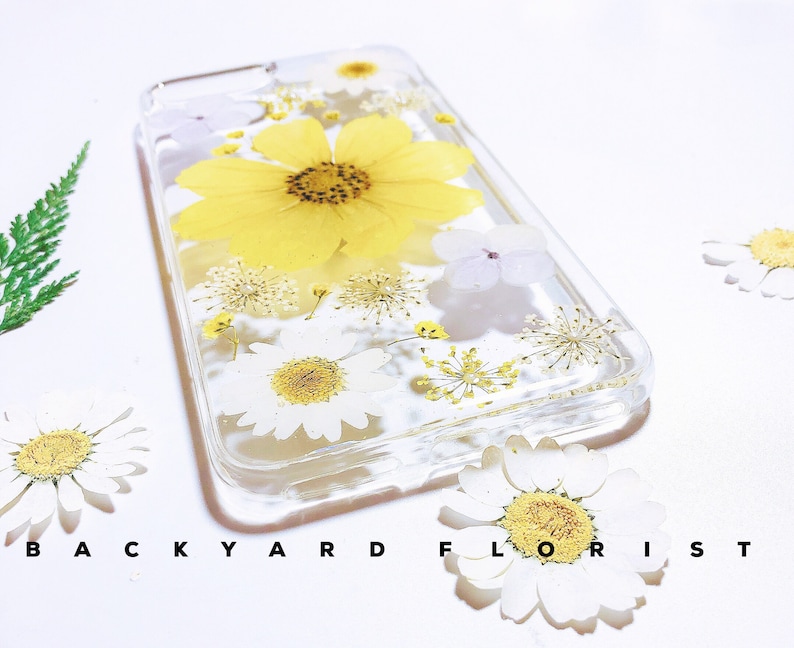 Peut inclure: Une coque de t&eacute;l&eacute;phone transparente avec des fleurs press&eacute;es. La coque pr&eacute;sente une grande fleur jaune avec des p&eacute;tales blancs et des fleurs blanches plus petites. La coque est &eacute;tiquet&eacute;e "BACKYARD FLORIST".