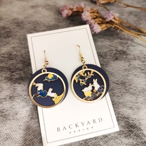 Puede incluir: Un par de pendientes colgantes de oro con un diseño de esmalte azul y blanco. Los pendientes presentan un círculo con un diseño de conejo y luna en un lado y un diseño de conejo y árbol en el otro. Los pendientes están en una tarjeta blanca con el texto "BACKYARD DESIGN".