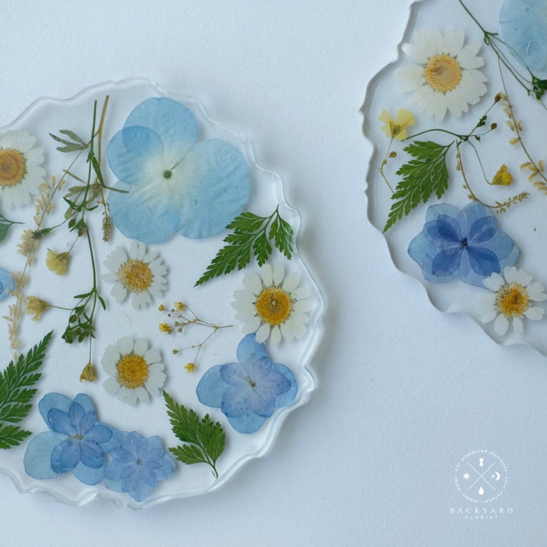 May include: Two round resin coasters with a white background and pressed flowers. One coaster features blue and white flowers, while the other features blue, white, and yellow flowers.