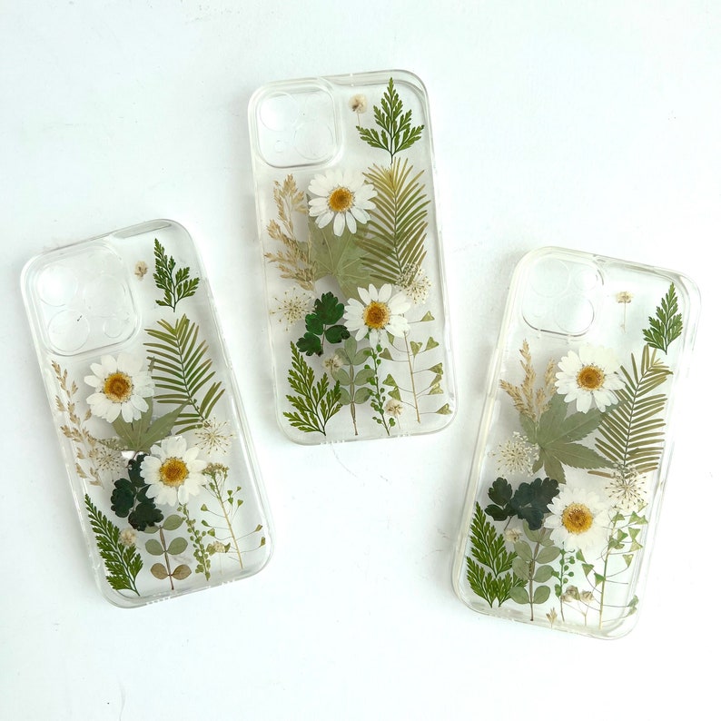 May include: Three clear phone cases featuring pressed white flowers and green leaves. The cases are designed for a smartphone with a rectangular shape.