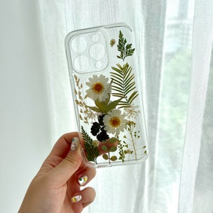 May include: A clear phone case featuring pressed white flowers and green leaves. The case is designed for a smartphone with a camera lens.