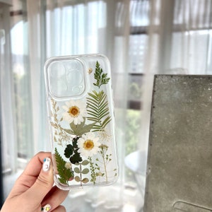 May include: A clear phone case with pressed white flowers and green leaves. The case is designed for an iPhone.