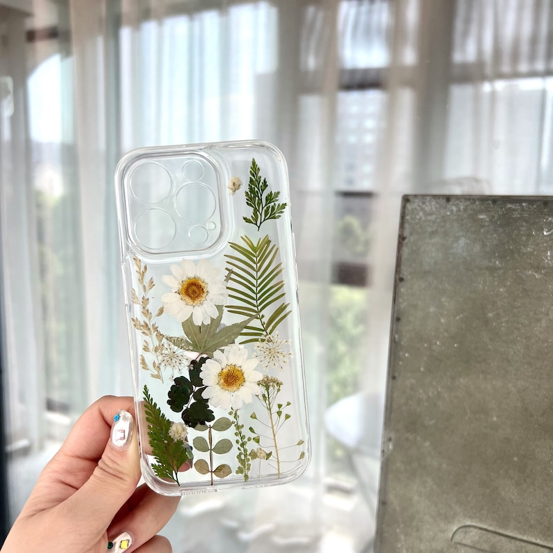 May include: A clear phone case with pressed white flowers and green leaves. The case is designed for an iPhone.