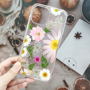 May include: Clear phone case with pressed flowers and greenery. The flowers are pink, white, and yellow. The case has a black logo that says "Backyard Florist" and "For treasured moments - soul, water, jewel, sun".
