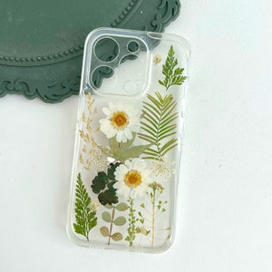 May include: A clear phone case with pressed white flowers and green leaves. The case is designed for an iPhone.