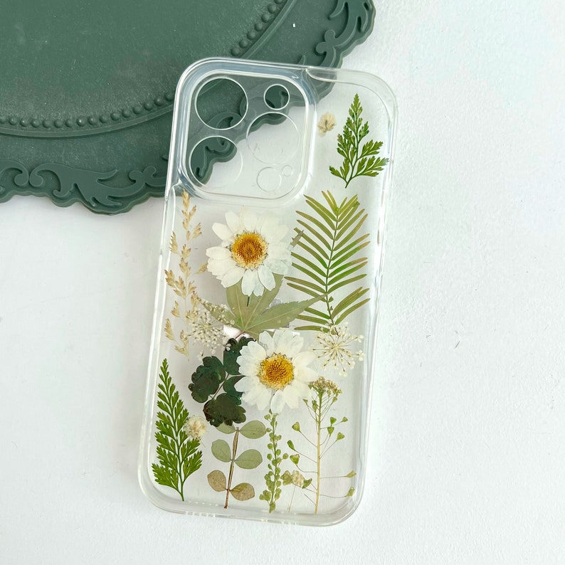 May include: A clear phone case with pressed white flowers and green leaves. The case is designed for an iPhone.