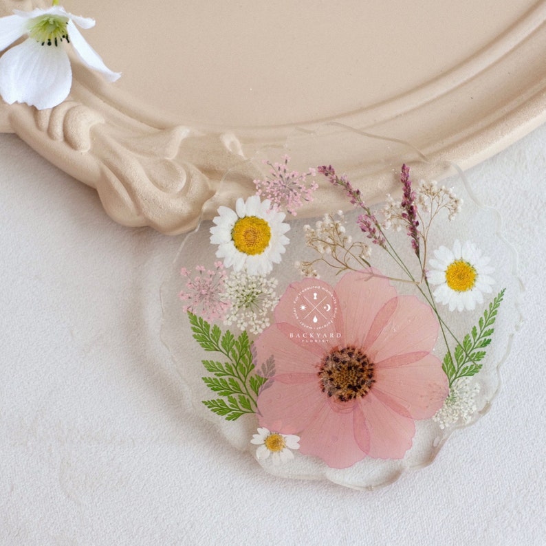 May include: A heart-shaped resin coaster with pressed flowers. The coaster features a pink flower in the center, surrounded by white daisies, small white flowers, and green ferns. The coaster has a white background and the text "BACKYARD" in black.