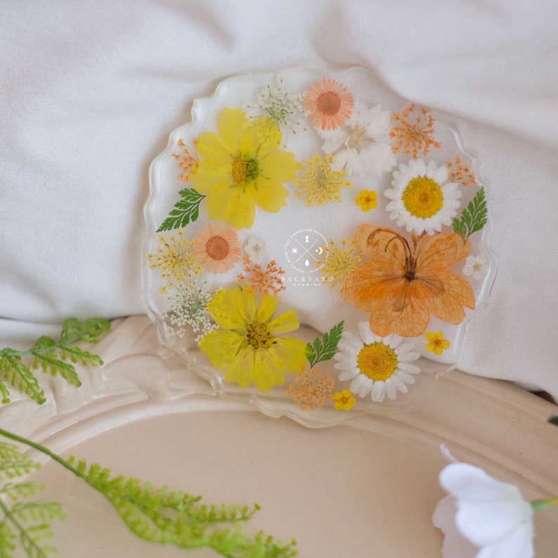 May include: A round, clear resin coaster with a pressed flower design. The coaster features a variety of yellow, orange, and white flowers, including daisies, cosmos, and a butterfly.