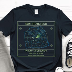May include: Navy blue t-shirt with a graphic featuring a radar-like map of San Francisco. Text includes "SAN FRANCISCO" and "PARKING ENFORCEMENT REAL-TIME UPDATES SOURCE UNDER MAINTENANCE". The shirt is paired with gray jeans and black sneakers.
