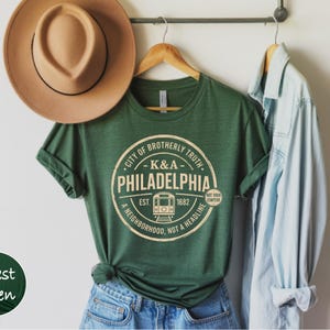 May include: Forest green t-shirt with a circular graphic that reads "City of Brotherly Truth, K&A Philadelphia, Est. 1682, A Neighborhood, Not a Headline." A tan hat hangs above the shirt. A light blue denim shirt hangs to the right.