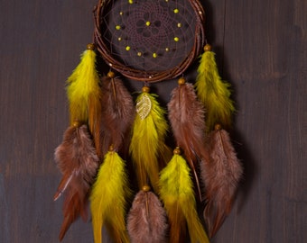 May include: A brown dreamcatcher with yellow and brown feathers hanging from it. The dreamcatcher is made of twigs and has a beaded web. The dreamcatcher is hanging against a dark brown wooden background. The text "Runiversia" is written below the dreamcatcher.