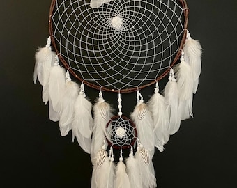 Puede incluir: Un atrapasueños blanco con aros de madera marrón y plumas blancas. El atrapasueños tiene un aro más pequeño con cuentas blancas y plumas que cuelgan de él.