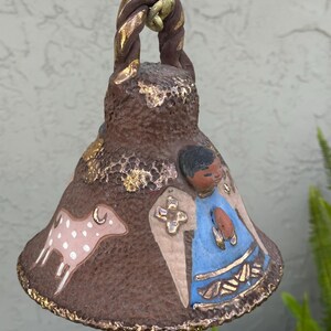 May include: A handcrafted ceramic bell with a brown textured finish, adorned with gold accents. The bell features a painted design of a figure in blue robes, a lamb, and a cross. It hangs from a gold-colored chain.