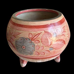 May include: A round, hand-painted ceramic pot with three small feet. The pot features a beige base with red and pink floral designs and stripes. A large gray flower is painted on the side.