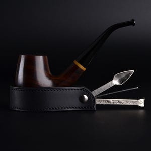 Puede incluir: Una pipa de tabaco de madera marrón oscuro con boquilla negra y una banda amarilla, apoyada detrás de un estuche de cuero negro que contiene tres herramientas de metal plateado para pipa, incluyendo un atacador, un escariador y un punzón.