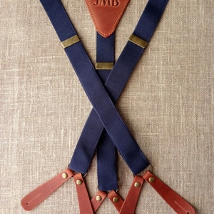 May include: Navy blue suspenders with brown leather accents. The leather patch at the back features the initials "JMB". The suspenders have brass-colored hardware and brown leather ends.