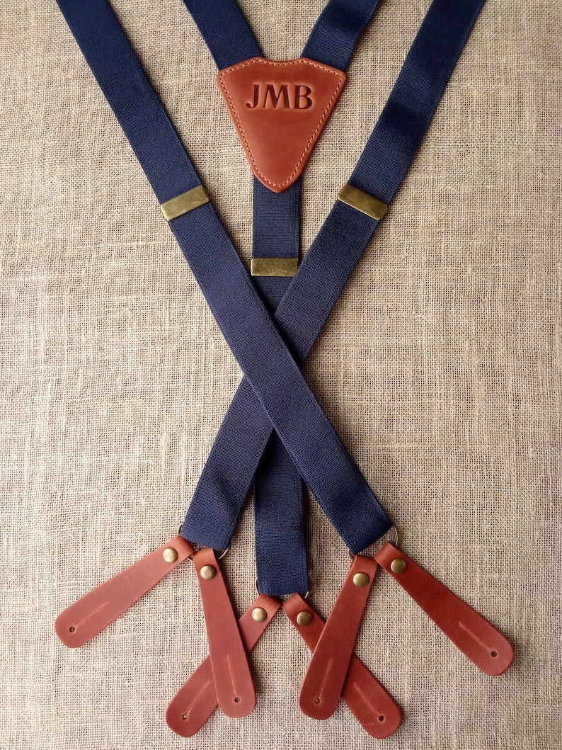 May include: Navy blue suspenders with brown leather accents. The leather patch at the back features the initials "JMB". The suspenders have brass-colored hardware and brown leather ends.