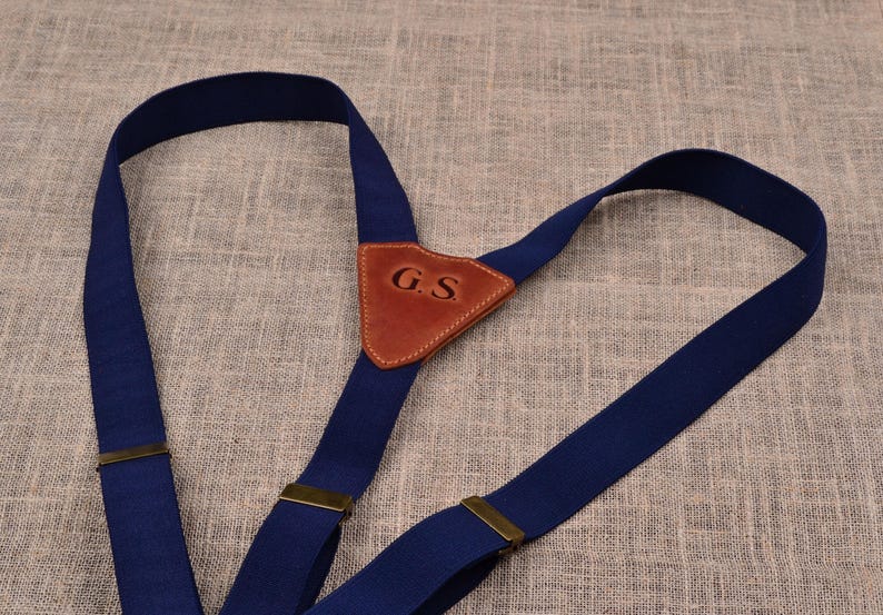 May include: Navy blue suspenders with a brown leather patch featuring the initials "G.S." The suspenders have brass-colored metal adjusters and are made of a sturdy, woven material. The design is classic and suitable for various outfits.