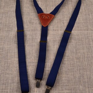 May include: Navy blue suspenders with metal clips and a brown leather triangle with the initials "G.S." These suspenders are a classic accessory for trousers, offering both style and functionality. The suspenders are made of a durable fabric.
