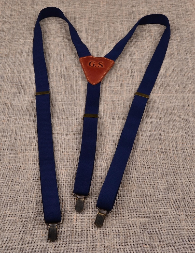 May include: Navy blue suspenders with metal clips and a brown leather triangle with the initials "G.S." These suspenders are a classic accessory for trousers, offering both style and functionality. The suspenders are made of a durable fabric.