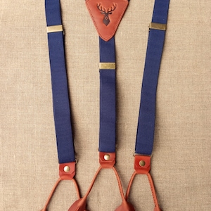 May include: Navy blue suspenders with brown leather accents. The suspenders feature a brown leather patch with a deer silhouette. The suspenders have brass-colored metal hardware and brown leather button attachments.
