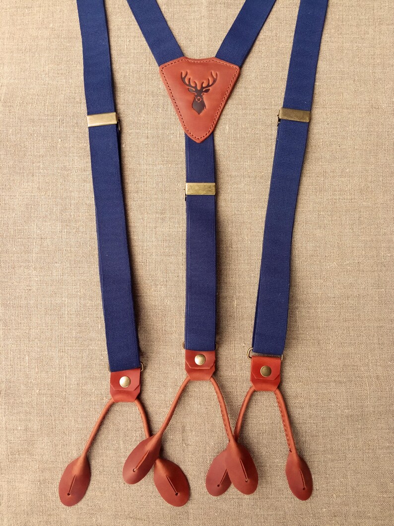 May include: Navy blue suspenders with brown leather accents. The suspenders feature a brown leather patch with a deer silhouette. The suspenders have brass-colored metal hardware and brown leather button attachments.