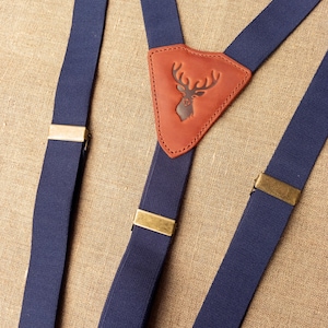 May include: Navy blue suspenders with brass-colored metal clasps. The center leather patch features a debossed deer head design. The suspenders are made of a sturdy, woven material.
