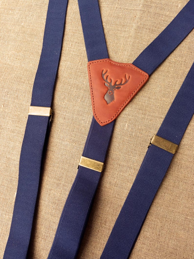 May include: Navy blue suspenders with brass-colored metal clasps. The center leather patch features a debossed deer head design. The suspenders are made of a sturdy, woven material.