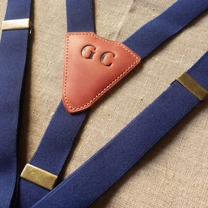 May include: Navy blue suspenders with brass-colored metal clasps and a brown leather back patch. The leather patch is embossed with the initials "GC". The suspenders are made of a sturdy, elastic material.