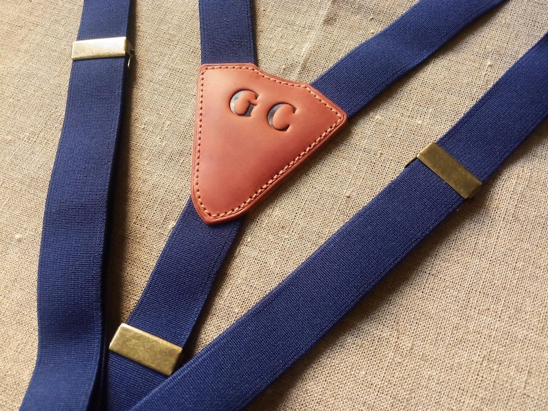 May include: Navy blue suspenders with brass-colored metal clasps and a brown leather back patch. The leather patch is embossed with the initials "GC". The suspenders are made of a sturdy, elastic material.