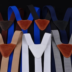 May include: Assortment of suspenders in various colors, including brown, blue, black, navy, white, and gray. Each pair features a leather or leather-like patch at the Y-back and adjustable straps. The white pair has "RAWS" embossed.