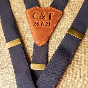 May include: Navy blue suspenders with brass-colored hardware and a brown leather patch. The leather patch is embossed with the initials "C&T" and the date "26.6.21". The suspenders are made of a woven material.